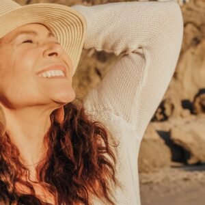 A joyful woman with a hat basks in the sunlight by the seaside, symbolizing relaxation and happiness.